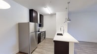This single-floor space showcases a contemporary kitchen designed with both style and functionality in mind. The kitchen is anchored by a galley-style layout along one wall, featuring dark espresso cabinetry that extends to the ceiling, providing ample storage. Stainless steel appliances, including a full-size refrigerator and a range with an overhead microwave, complement the modern aesthetic.

A standout feature is the white quartz peninsula countertop, which extends outward to create additional workspace and a casual dining or gathering area. An undermount sink with a sleek chrome faucet is integrated into the peninsula, and two polished chrome industrial-style pendant lights hang above, adding a stylish focal point.

The light wood-tone flooring contrasts elegantly with the dark cabinetry, lending warmth to the space. Bright white walls and recessed fluorescent ceiling lighting keep the kitchen feeling open and well-illuminated. The clean lines, neutral palette, and quality finishes throughout create a cohesive and inviting environment suited for everyday living.