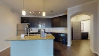 This single-story residence showcases a cohesive modern design carried consistently throughout its rooms. The kitchen serves as a central highlight, featuring dark espresso cabinetry that extends to the ceiling, providing generous storage. A large granite-topped island with an integrated sink anchors the space, illuminated by pendant lights and a track lighting system. White subway tile backsplash contrasts sharply against the dark cabinetry, while stainless steel appliances, including a refrigerator, range, and built-in microwave, complete the setup. An elegant arched doorway leads further into the unit. Dark hardwood-style flooring runs continuously throughout, grounding every room with a warm, rich tone. The living area is open and generously proportioned, benefiting from natural light through blinds-fitted windows that offer outdoor views. Bedrooms are well-sized with neutral walls and ceiling fans. A hallway connects the bedroom to a closet and bathroom, which features a dark-cabineted vanity with a granite countertop and a grab bar. A separate bathroom includes a vanity with matching granite and access to an in-unit laundry area. Crown molding accents the kitchen and common areas, adding a refined architectural touch throughout.