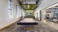 Located at 350 E Congress St in downtown Tucson, Arizona, this contemporary game and lounge room offers a well-appointed communal amenity space designed for entertainment and social interaction. The room is anchored by a sleek, modern pool table with a gold-toned base and tan felt surface, positioned beneath an eye-catching cluster pendant light that draws attention to the boldly painted olive-green accent ceiling above. A wall-mounted large-screen television is prominently displayed against the olive-green feature wall, creating a focal point for the space. Additional game tables, including a shuffleboard and foosball table, are positioned throughout the room, offering varied entertainment options. The flooring features a textured, patterned carpet with a stone-like aesthetic, complemented by polished concrete in the adjacent corridor areas. Exposed concrete structural columns and visible ceiling infrastructure lend an urban industrial character, while recessed lighting ensures the space is well-lit. Framed artwork adorns the white walls, adding a curated aesthetic touch. The room connects openly to a broader lounge area, visible through large black-framed glass partitions, extending the sense of space and allowing natural light to filter through from floor-to-ceiling windows beyond.