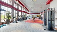 Located at 20200 Poplar Creek Pkwy in Brookfield, Wisconsin, this expansive fitness center spans a generous single floor and is divided into two dedicated exercise rooms, offering a comprehensive and well-equipped workout environment. The facility is defined by its striking architectural design, featuring soaring ceilings, bold red accent beams, and large wood-framed windows that flood the interior with natural light and provide open views of the surrounding exterior. The main exercise room is outfitted with a wide array of strength-training machines, free weights, adjustable benches, and a full dumbbell rack, all set on vibrant red rubber flooring that contrasts with the neutral carpeted zones. Pendant lighting and ceiling fans add both functional illumination and comfort throughout the space. The second exercise room features a distinctly curved wall of floor-to-ceiling windows, creating a bright, airy atmosphere ideal for cycling classes. It is equipped with a row of stationary spin bikes and a checkered red-and-grey tile floor that adds a dynamic visual element. Additional amenities include a functional training rig with TRX straps and resistance bands, medicine balls, stability balls, and a line of cardio machines including ellipticals and stair climbers. Warm wood-framed glass doors connect the fitness center to adjacent amenity spaces, contributing to a cohesive and polished overall design.