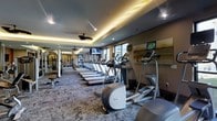 This modern fitness center offers a comprehensive workout environment designed to accommodate a wide range of exercise needs. The space is anchored by an impressive row of Precor cardio machines, including treadmills and ellipticals, all aligned to take advantage of natural light streaming in through a series of windows along the wall. Mounted flat-screen televisions are positioned throughout, allowing members to stay entertained during workouts. The facility extends generously, with a strength and free weights area visible in the background, outfitted with various resistance machines and equipment. The interior design strikes a balance between functionality and style, featuring a tray ceiling with warm cove lighting that creates an inviting ambiance, complemented by recessed lighting and ceiling fans that ensure comfort throughout. The flooring consists of durable, patterned carpet suited for a high-traffic fitness environment. Large mirrors are strategically placed to enhance the sense of space and allow users to monitor their form. The overall atmosphere is clean, modern, and energizing — well-suited for both casual exercisers and dedicated fitness enthusiasts.