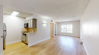 Located on Mark Dabling Boulevard in Colorado Springs, Colorado, this single-floor unit offers a well-designed open-concept layout that seamlessly connects the kitchen and living areas. Light hardwood flooring runs throughout, contributing to a clean, airy aesthetic that is further enhanced by white walls and a textured ceiling.

The kitchen is positioned along one side of the open space, featuring granite countertops, gray cabinetry, a tile backsplash, a built-in electric cooktop, an over-the-range microwave, and a dishwasher. A raised countertop edge provides a casual bar-style seating option and serves as a natural visual divider between the kitchen and the main living area.

The living area is generously proportioned, bathed in natural light from a large multi-pane window that overlooks the exterior. Wall-mounted sconce lighting adds additional ambiance throughout the shared space. A separate room is accessible via an interior doorway, offering additional functional space.

The bathroom includes a full tub and shower combination with white tile surround, a grab bar, granite-topped vanity, and tile flooring, maintaining a consistent, clean finish throughout the unit.
