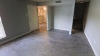 This two-floor home offers a clean, functional layout with a cohesive design throughout. The main living area is finished with durable wood-look plank flooring in a warm gray-brown tone, complemented by neutral gray walls and crisp white baseboards that create a modern, understated aesthetic. A wide open archway connects the living space to an adjacent room — likely a dining or kitchen area — where a ceiling fan with a light fixture provides both air circulation and illumination. The connected space features lighter flooring and a countertop or bar area visible in the background, suggesting a kitchen or breakfast nook. An entry door is conveniently located in the main living area, offering direct access to the space. Overhead, a ceiling-mounted HVAC vent ensures year-round climate control. A staircase is partially visible at the edge of the room, leading to the second floor and hinting at additional living space above. The overall ambiance is bright, neutral, and move-in ready, with a simple, practical design that lends itself well to personalization. The open sightlines between rooms enhance the sense of flow and connectivity throughout the home.