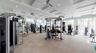 This spacious fitness center offers a full suite of amenities designed to support a wide variety of workout routines. The facility is thoughtfully organized into distinct zones, allowing members to move seamlessly between cardio, strength training, and functional fitness areas. A row of modern treadmills and an elliptical machine face expansive floor-to-ceiling windows, framing lush outdoor views of palm trees and the surrounding community, creating an energizing atmosphere during workouts. The strength training zone is outfitted with a multi-functional cable rack, a heavy punching bag, TRX suspension trainers, and a full complement of selectorized weight machines, all reflected in large wall-mounted mirrors that amplify the sense of space. Rowing machines and recumbent bikes occupy a dedicated area alongside a wall-mounted flat-screen television. A functional training station features stability balls displayed on a freestanding rack, adding a versatile element to the floor plan. The architectural design showcases a coffered ceiling with recessed lighting and ceiling fans, contributing to both the polished aesthetic and comfortable climate control. Neutral-toned carpet tile flooring and soft sage-green walls lend a clean, modern feel throughout. Glass partitions connect the fitness center to adjacent lounge areas, maintaining an open and airy environment.