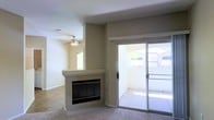 This single-floor residence offers a well-designed open layout that maximizes both comfort and functionality. The main living area is a welcoming space anchored by a corner fireplace with a granite-style surround, providing a natural focal point for the room. Neutral-toned walls and light carpet flooring create a clean, versatile backdrop throughout the space. A ceiling fan overhead contributes to year-round comfort while adding a practical architectural detail to the living area. Natural light flows generously into the interior through multiple windows and a large sliding glass door, which opens directly onto a private outdoor patio. The patio extends the living space outdoors, offering views of surrounding greenery and neighboring structures. The transition between indoor and outdoor areas is seamless, making the patio a natural extension of the interior. The open flow between spaces enhances the sense of roominess throughout. Overall, the residence presents a straightforward and functional design, with thoughtful touches such as the fireplace and abundant natural light contributing to a comfortable and inviting ambiance suited for everyday living.