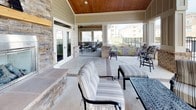 This expansive covered outdoor patio is thoughtfully designed for both relaxation and socializing. The ceiling is finished with warm wood paneling, adding a refined, lodge-inspired aesthetic that contrasts beautifully with the structural columns clad in stacked stone and brick. A prominent floor-to-ceiling stone fireplace serves as a central architectural focal point, complete with a wood mantle and a glass-enclosed gas firebox, offering a cozy gathering spot during cooler evenings. Brick-inlaid pathways provide subtle definition to the concrete patio surface, adding texture and visual interest underfoot. Multiple seating arrangements are spread throughout the space, including cushioned sofas, lounge chairs, and cast-iron accent tables, accommodating a variety of social configurations. White French doors connect the covered patio to an interior space, ensuring seamless indoor-outdoor flow. At the open edge of the patio, wrought iron fencing frames direct views of a community swimming pool, with manicured landscaping and multi-story residential buildings visible beyond. Ceiling fans are installed throughout, enhancing comfort during warmer seasons. Together, these elements create a well-appointed outdoor living area suited for year-round use.