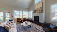Located at Wareham Road in Plymouth, Massachusetts, this single-floor community clubhouse is thoughtfully designed to offer residents a range of social, recreational, and wellness amenities. The main lounge area immediately establishes a warm, inviting atmosphere with soaring vaulted ceilings clad in shiplap and a striking floor-to-ceiling tiled fireplace as the centerpiece. Light hardwood flooring runs throughout, complementing the neutral palette and curated furnishings. A row of large windows with plantation shutters floods the space with natural light while offering views of the surrounding grounds. The seating area features a comfortable arrangement of sofas and leather accent chairs, making it an ideal gathering space. Adjacent to the lounge, the game room continues the vaulted ceiling design and includes a full-size billiards table, a built-in shelving unit with a mounted flat-screen television, and a cozy nook with bench seating — all set against a textured accent wall. The fitness center is generously sized and well-equipped with cardio machines including treadmills, ellipticals, and steppers, alongside an array of exercise balls, resistance equipment, and floor mats. Full-length mirrors and vibrant wall art energize the space. Together, these areas create a cohesive, modern clubhouse environment designed for both relaxation and an active lifestyle.