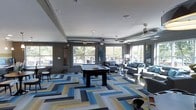 Located at 811 Spring Forest Road in Raleigh, NC, this single-floor community clubhouse offers a well-appointed and vibrant gathering space designed for both recreation and relaxation. The open layout is anchored by a full-size pool table positioned at the center of the room, making it an immediate focal point upon entry. A bold, geometric-patterned carpet in shades of blue, gold, gray, and white adds a striking visual element underfoot, lending the space a contemporary and energetic aesthetic. The lounge area is thoughtfully furnished with plush gray sofas and modern accent tables, creating a comfortable setting for socializing. Exposed ductwork runs along the ceiling, complementing the industrial-chic design sensibility, while ceiling fans ensure year-round comfort. Large windows line multiple walls, flooding the interior with generous natural light and offering views of the surrounding greenery outside. The gray wall palette provides a neutral yet sophisticated backdrop that ties together the colorful furnishings and décor. Overall, the space strikes a balance between lively entertainment and relaxed comfort, making it an ideal amenity hub for the community it serves.