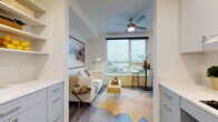 This thoughtfully designed single-floor unit maximizes every inch of its layout through a clean, contemporary aesthetic anchored by neutral tones and wood-look flooring throughout. The main living and sleeping area serves as the heart of the space, highlighted by large corner windows that flood the room with natural light and frame elevated views of the surrounding neighborhood and landscape. A ceiling fan overhead adds practical comfort to the airy atmosphere.

The sleeping area is well-proportioned, accommodating a full-sized bed alongside a stylish bistro-style table and stools positioned directly beside the windows — an ideal spot to enjoy the panoramic outlook. A built-in nook provides open shelving and storage space, while a doorway connects directly to the bathroom. The bathroom is clean and minimal, featuring chrome fixtures and efficient use of space.

A separate living nook, framed by white cabinetry on either side, offers a cozy retreat with room for seating and a small accent table, further benefiting from the abundant natural light. The galley-style kitchen runs along the entry corridor, lined with white cabinetry, light countertops, a single-basin sink, and a full-sized refrigerator — offering a complete and functional cooking space within a compact footprint.