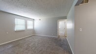 This single-story home offers three bedrooms, one bathroom, a kitchen, and a hallway, all arranged within a compact yet functional floor plan. The generously sized primary bedroom features neutral gray walls, wall-to-wall carpeting, a textured ceiling, and windows fitted with blinds that allow natural light to filter through. A doorway from the primary bedroom connects directly to the bathroom, adding convenience. The secondary bedrooms are similarly finished with carpet, neutral-toned walls, textured ceilings, and individual closets equipped with wire shelving for organized storage. Each bedroom includes at least one window with blinds. The kitchen is outfitted with light wood cabinetry, granite-style laminate countertops, a double stainless steel sink, and a dishwasher, providing practical functionality. The bathroom features a full tub-shower combination with white tile surround, a toilet, and wood-look vinyl flooring. The main living area is carpeted with a textured ceiling and includes a front entry door, offering a straightforward open layout. Throughout the home, consistent gray wall tones, white trim, and carpet flooring create a cohesive, neutral aesthetic.