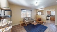This single-floor home offers a thoughtfully arranged layout that balances comfort and functionality throughout. The living room serves as the heart of the space, featuring warm hardwood-style flooring, neutral beige walls, and a large window fitted with horizontal blinds that invite an abundance of natural light. The room flows seamlessly toward the kitchen, which is visible through an open passageway and is equipped with rich warm-toned wood cabinetry, dark granite countertops, a stainless steel double sink, and a full suite of stainless steel appliances including a range, built-in microwave, and dishwasher. Tile flooring completes the kitchen's practical yet polished aesthetic. The bedrooms include a carpeted room with multiple windows on two walls, providing excellent natural light and ventilation, as well as a cozier bedroom with a closet and a single window. The bathroom is a standout feature, showcasing large-format dark tile surround walls, a soaking tub with shower, a glass block window for privacy and diffused light, and tile flooring that carries a cohesive, modern feel throughout. The overall ambiance of the home is warm, functional, and move-in ready.