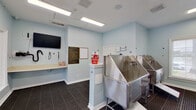 This single-floor pet spa is a purpose-built grooming facility designed with both functionality and charm in mind. The space is finished in a soothing light blue palette throughout, complemented by crisp white trim and accents that create a clean, welcoming atmosphere.

The main grooming area is the centerpiece of the facility, housing multiple professional-grade stainless steel wash tubs equipped with integrated sprayer attachments. The tubs feature angled entry ramps for easy pet access and are arranged to accommodate simultaneous use. Anti-fatigue, non-slip rubber matting covers the floors throughout the grooming zones, prioritizing safety for both pets and handlers.

The facility includes a dedicated counter area with storage, a wall-mounted flat-screen television, and posted Pet Spa Rules signage to guide users. Playful paw print wall decals add a lighthearted touch to the décor, reinforcing the pet-friendly nature of the space. A separate room with wood-look flooring and additional paw print accents offers a flexible utility area.

Natural light enters through large windows that frame views of the surrounding community, while recessed ceiling speakers and flush-mounted fluorescent lighting ensure a bright, well-lit environment. The facility also features direct exterior access through glass-paneled doors, making entry and exit convenient for pets and their owners alike.