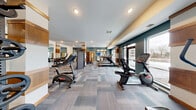 Located at 9900 56th Ave N in Plymouth, Minnesota, this fitness center offers a spacious and thoughtfully designed workout environment across two exercise rooms on a single floor. The facility is finished with a neutral, multi-toned carpet in a herringbone-inspired pattern that adds a modern aesthetic throughout. Architectural columns accented with warm wood paneling provide visual structure and complement the teal and white color palette of the walls, creating an inviting and energetic atmosphere.

The main exercise area is generously sized and bathed in natural light from large floor-to-ceiling windows and glass doors, offering views of the surrounding outdoor landscape. Cardio equipment includes treadmills, recumbent bikes, and upright cycles, while strength training needs are met with a power rack with free weights and a multi-function cable machine. A dedicated dumbbell rack with wall-mounted mirrors lines one side of the space, supporting a variety of free weight exercises.

A separate, glass-partitioned room provides an enclosed area for focused workouts or stretching, featuring hard flooring for versatility. Recessed lighting throughout ensures consistent illumination, complementing the ample natural light. Built-in shelving and wall art add a polished, community-oriented finish to the space, making this fitness center both functional and aesthetically refined.