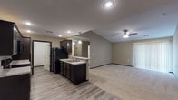 This single-story unit is designed with an open-concept layout that connects the kitchen and living areas seamlessly. The kitchen is well-appointed with dark espresso cabinetry, granite-style countertops, black appliances including a refrigerator, range, and microwave, and light wood-look vinyl flooring. A peninsula with a double sink and pendant lighting overhead serves as a natural divider between the kitchen and the adjoining living space.

The living area features vaulted ceilings, a ceiling fan with integrated lighting, and plush neutral carpet, creating a comfortable and spacious atmosphere. Large vertical-blind-covered sliding doors allow generous natural light to fill the room. Additional bedrooms are carpeted and equipped with ceiling fans, offering well-sized, functional spaces with windows providing natural light.

Two bathrooms are present: one featuring a dark vanity with granite-style countertops, a large mirror, and a tub-shower combination, and a second, more expansive bathroom with a wide vanity, wood-look flooring, and ample open floor space. Throughout, neutral taupe wall tones, dark trim accents, and recessed lighting contribute to a cohesive and modern aesthetic.