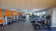 Located at 1237 Enclave Cir in Arlington, Texas, this well-appointed fitness center offers a comprehensive workout environment across a single floor. The facility is characterized by its bold orange and gray color scheme, corrugated metal accent walls, and elevated ceilings that lend an industrial yet energetic aesthetic throughout.

The main exercise floor is generously sized and outfitted with a extensive row of SportsArt cardio machines, including treadmills and ellipticals, positioned along floor-to-ceiling mirror walls that amplify the sense of space. Strength training equipment and multi-use cable machines occupy a dedicated section of the floor, all finished with durable dark rubber flooring suited for high-traffic use.

A separate studio or functional training area features light hardwood flooring and is equipped with stability balls, medicine balls, dumbbells, and wall mirrors — ideal for stretching, group classes, or individual training sessions. Natural light filters in through multiple windows, offering outdoor views of the surrounding greenery and pool area.

The facility also includes a restroom, wall-mounted amenity stations, and direct glass door access to the outdoor pool area, creating a seamless connection between indoor and outdoor fitness spaces. The overall ambiance is modern, energetic, and functional, designed to support a full range of fitness activities.