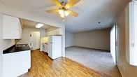 This single-floor space offers an open-concept design that seamlessly connects the kitchen and living areas. The kitchen is equipped with white cabinetry, a white refrigerator, and a gas range, complemented by a fluorescent overhead light that brightens the workspace. White upper cabinets provide ample storage, while the layout keeps the cooking area functional and accessible. The transition from the kitchen to the living area is marked by a shift from warm hardwood-style flooring to neutral carpet, defining each zone within the open plan. The living area is spacious, offering room for a variety of furniture arrangements, and is centered by a ceiling fan with an integrated light fixture, ensuring both air circulation and illumination. The neutral gray walls throughout provide a versatile backdrop suitable for various interior styles. A hallway near the kitchen leads to additional areas of the unit, including what appears to be a utility or storage space. Overall, the layout is practical and straightforward, making efficient use of the available space.