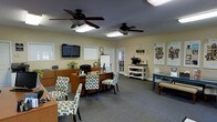 This single-story office space offers a functional and inviting environment designed to accommodate both staff and visiting clients. The open main area is carpeted in a neutral gray tone and features dual ceiling fans that contribute to a comfortable atmosphere throughout the day. Natural light filters in through multiple windows, complementing the warm, beige-toned walls and giving the space an approachable feel.

The central work area includes a wood-toned desk station equipped with a computer monitor and printer, providing a practical hub for daily operations. Several patterned accent chairs in a green-and-white geometric print are arranged throughout the space, offering comfortable seating for clients. A wall-mounted flat-screen television is positioned for easy viewing, while a whiteboard nearby supports presentations or collaborative discussions.

A dedicated refreshment station along one wall features a coffee setup, adding a hospitable touch for guests. Informational displays and framed materials are mounted on the walls, reinforcing the professional character of the space. A rear door with glass panel inserts adds architectural interest and allows additional light into the interior.