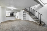 This two-floor space offers a well-organized layout with functional design throughout. The main level centers around a generously sized living area finished with plush carpet, highlighted by a striking fireplace framed in board-and-batten surround with a raised brick hearth and glass-panel doors. A staircase with sleek black metal railings and carpeted treads rises prominently alongside the living area, connecting to the upper level. The kitchen is equipped with gray cabinetry, dark countertops, a peninsula bar, and black appliances including a range and refrigerator, with a window providing natural light. A bathroom on the main level features dark wood vanity cabinetry, light countertops, and wood-look vinyl flooring. Upstairs, an open loft overlooks the main level through matching black metal railings and is fitted with a ceiling fan, offering a versatile, light-filled space. A spacious walk-in closet with dual-level hanging rods and carpet flooring provides generous storage. Throughout, textured ceilings add dimension, and neutral tones create a clean, cohesive aesthetic across both floors.