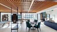 This thoughtfully designed single-floor amenity space blends sophisticated style with a range of recreational and social features. A striking wood slat ceiling runs through the games area, where a full-size pool table and shuffleboard table take center stage beneath integrated lighting. Large industrial-style grid windows flood the space with natural light and offer views of the surrounding cityscape and outdoor pool deck.

The lounge area is anchored by plush navy sectional sofas and accent chairs arranged on a vibrant teal area rug, creating a welcoming and cohesive social environment. Open shelving with warm wood tones and dark metal frames adds architectural interest throughout, while framed artwork and curated décor lend a gallery-like quality to the walls.

A cozy seating nook near a sleek modern fireplace provides an intimate retreat, accented by an eye-catching mosaic tile surround in rich greens and blues and a decorative log display. Direct access to the outdoor pool deck is available through floor-to-ceiling glass doors, connecting the interior effortlessly to the exterior amenity space. The light-toned tile flooring, high ceilings in select areas, and cohesive blue and wood color palette unify the space into a refined, resort-inspired environment suited for relaxation and entertainment alike.