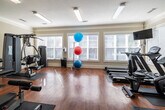 This fitness center is a comprehensively outfitted exercise facility designed to support a wide range of workout routines. The space is laid out across a single floor and features warm hardwood flooring throughout, complemented by neutral-toned walls, crown molding, and a textured ceiling with flush-mounted fluorescent lighting that ensures consistent illumination across the room.

Natural light flows generously through multiple large windows fitted with white horizontal blinds, creating a bright and energizing atmosphere. The cardio zone is well-stocked with treadmills, elliptical trainers, and a stationary bike, all arranged along the windowed walls and accompanied by a wall-mounted flat-screen television for entertainment during workouts.

The strength training area features a multi-station cable machine, several selectorized weight machines including a Body-Solid unit, a dedicated free weights rack with dumbbells, and an adjustable workout bench. Stability balls in multiple sizes add versatility for core and balance training. Protective rubber floor mats are placed beneath equipment throughout the space.

An elegant set of French doors with glass panels provides access to the room, adding a refined architectural touch. A whiteboard and informational charts on the wall offer workout guidance. The overall ambiance is clean, functional, and welcoming, making it a practical fitness destination.