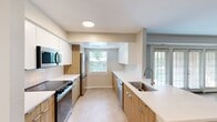 This 720-square-foot, one-bedroom, one-bathroom apartment at 8800 Queensmere Place in Richmond, Virginia offers a well-designed single-floor layout. The galley-style kitchen is a standout feature, showcasing contemporary two-tone cabinetry with warm wood lower cabinets and crisp white upper cabinets, paired with white quartz countertops and stainless steel appliances including a refrigerator, dishwasher, microwave, and range. Natural light fills the kitchen through multiple windows overlooking greenery. The open living room is finished with rich hardwood flooring and features a set of large glass patio doors that bring in abundant natural light and provide outdoor access. The 200-square-foot primary bedroom is carpeted for comfort and includes two closets—one a bifold-door closet and one additional storage closet—plus a window with natural views. The bathroom is clean and functional, featuring a full tub/shower combo with white tile surround, a vanity with mirror, and tile flooring. Neutral wall tones throughout the unit create a calm, cohesive ambiance well-suited for a variety of interior styles.