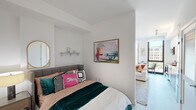 This single-floor residence is thoughtfully laid out to maximize both functionality and style. The living area serves as the heart of the space, featuring crisp white walls, light hardwood-style flooring, and an open layout that promotes a comfortable, airy atmosphere. A low-profile media console anchors the room alongside a curated gallery wall, a statement fiddle-leaf fig plant, and a plush white sofa accented with warm-toned throw pillows. Natural light flows generously through floor-to-ceiling windows, offering views of the surrounding cityscape.

The bedroom is a cozy yet well-designed retreat, characterized by clean white walls, a tufted upholstered headboard, and vibrant, eclectic bedding that adds personality to the space. A gold-framed oval mirror, a decorative wall shelf, and framed abstract artwork complete the room's aesthetic. An open sightline connects the bedroom to the living area, enhancing the sense of flow throughout the unit.

The bathroom features large-format neutral tile flooring, a sleek wood-toned vanity with an integrated sink, and crisp white walls. Thoughtful decorative accents, including framed artwork and a modular wall shelf with greenery, give the bathroom a polished, well-considered finish.