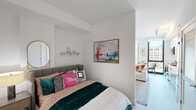 This single-floor residence is thoughtfully laid out to maximize both functionality and style. The living area serves as the heart of the space, featuring crisp white walls, light hardwood-style flooring, and an open layout that promotes a comfortable, airy atmosphere. A low-profile media console anchors the room alongside a curated gallery wall, a statement fiddle-leaf fig plant, and a plush white sofa accented with warm-toned throw pillows. Natural light flows generously through floor-to-ceiling windows, offering views of the surrounding cityscape.

The bedroom is a cozy yet well-designed retreat, characterized by clean white walls, a tufted upholstered headboard, and vibrant, eclectic bedding that adds personality to the space. A gold-framed oval mirror, a decorative wall shelf, and framed abstract artwork complete the room's aesthetic. An open sightline connects the bedroom to the living area, enhancing the sense of flow throughout the unit.

The bathroom features large-format neutral tile flooring, a sleek wood-toned vanity with an integrated sink, and crisp white walls. Thoughtful decorative accents, including framed artwork and a modular wall shelf with greenery, give the bathroom a polished, well-considered finish.