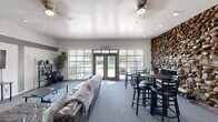 This single-floor space functions as a well-appointed community or leasing clubhouse, designed to accommodate both social gatherings and administrative functions. The layout is open and generously proportioned, with gray carpet flowing throughout the main areas and recessed lighting complementing ceiling fans overhead.

The most striking architectural feature is a floor-to-ceiling stacked stone accent wall, which anchors the dining area and adds a bold, rustic contrast to the otherwise clean, neutral interior. Adjacent to the stone wall, high-top tables with black bar stools provide a casual seating area ideal for meetings or informal gatherings.

A lounge area furnished with leather sofas and a coffee table offers a comfortable retreat, while a wall-mounted flat-screen television and media console make the space suitable for presentations or entertainment. The kitchenette, visible from the main area through an open pass-through, includes gray cabinetry and standard appliances.

Natural light enters generously through large windows fitted with vertical blinds and a set of glass French doors at the entry, which open to an outdoor view and are accented with a welcoming sign above. A small workstation near the entrance suggests a reception or leasing function. The overall ambiance is clean, modern, and inviting.