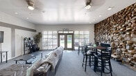 This single-floor space functions as a well-appointed community or leasing clubhouse, designed to accommodate both social gatherings and administrative functions. The layout is open and generously proportioned, with gray carpet flowing throughout the main areas and recessed lighting complementing ceiling fans overhead.

The most striking architectural feature is a floor-to-ceiling stacked stone accent wall, which anchors the dining area and adds a bold, rustic contrast to the otherwise clean, neutral interior. Adjacent to the stone wall, high-top tables with black bar stools provide a casual seating area ideal for meetings or informal gatherings.

A lounge area furnished with leather sofas and a coffee table offers a comfortable retreat, while a wall-mounted flat-screen television and media console make the space suitable for presentations or entertainment. The kitchenette, visible from the main area through an open pass-through, includes gray cabinetry and standard appliances.

Natural light enters generously through large windows fitted with vertical blinds and a set of glass French doors at the entry, which open to an outdoor view and are accented with a welcoming sign above. A small workstation near the entrance suggests a reception or leasing function. The overall ambiance is clean, modern, and inviting.