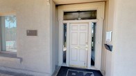 This single-floor residence makes a strong first impression with its classic exterior entry, featuring a six-panel white door framed by sidelights and a transom window above, all set within a stucco facade. A decorative bronze plaque accents the entryway, adding a touch of character and distinction.

Inside, the home is defined by warm hardwood flooring that flows throughout the main living areas, complemented by taupe-toned walls that create a cohesive and inviting atmosphere. The spacious living room serves as the heart of the home, anchored by a marble-surround fireplace with a wall-mounted television above. Vaulted ceilings and a chandelier lend an open, airy quality to the space, while an upright piano and an abundance of natural light streaming through vertical-blind windows add warmth and personality.

Adjacent seating areas provide versatile living arrangements. One casual sitting area features wicker chairs with teal cushions alongside a wall-mounted cue rack, suggesting a relaxed, leisure-oriented space. A secondary seating nook presents a pair of elegant tufted white armchairs positioned near a classic console table. Each area benefits from large windows that invite natural light and offer views of lush greenery outside, enhancing the overall sense of comfort and livability throughout the home.