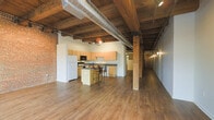 This single-floor loft space showcases a striking blend of industrial and modern design elements. The open-plan layout creates a generous, flowing interior that feels expansive and versatile. Overhead, heavy timber beams run across the ceiling alongside an exposed HVAC duct, lending the space a raw, warehouse-inspired aesthetic that is both functional and visually compelling. The original brick walls add warmth and texture, serving as a defining architectural feature throughout.

The kitchen is centrally positioned within the open floor plan, offering light maple cabinetry, dark countertops, and a full suite of appliances including a refrigerator and built-in microwave. A freestanding kitchen island with bar-height seating provides a casual dining and gathering area, making it ideal for both everyday use and entertaining.

Warm hardwood floors extend across the entire space, complementing the natural tones of the wood beams and cabinetry. Track lighting mounted to the beams ensures the space is well-lit while maintaining its industrial character. A long corridor leads to additional areas of the unit, accented with wall-mounted lighting that adds a stylish, gallery-like quality. Overall, the space delivers a cohesive, characterful environment with strong architectural bones and an inviting ambiance.