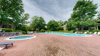 Located in the Birkdale Valley community of Charlotte, NC, this outdoor amenity area offers a well-appointed resort-style retreat designed for relaxation and leisure. The centerpiece is a generously sized, kidney-shaped swimming pool with clear turquoise water, complemented by a separate circular hot tub/spa positioned nearby on the expansive brick paver deck. The classic herringbone-patterned red brick hardscape extends throughout the space, providing ample room for lounging and movement. Rows of white and gray chaise lounge chairs are arranged along the pool's perimeter, offering plenty of seating for sunbathing or unwinding poolside. Mature trees frame the entire area, creating a lush, park-like canopy that provides both shade and a sense of seclusion. Neatly trimmed hedgerows and manicured landscaping add a polished, well-maintained aesthetic throughout. Stone retaining walls add a natural, architectural character to the grounds, while classic lamp posts contribute to the area's charm. A brick clubhouse building is visible in the background, enhancing the community feel. The overall ambiance is serene and inviting, blending classic architectural elements with abundant natural greenery for a comfortable outdoor living experience.