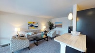 This single-floor residence showcases a thoughtfully designed open-concept layout that connects the living area to the kitchen, creating a fluid and functional living space. The living room is carpeted and comfortably sized, accommodating a tufted sofa, a pair of patterned accent chairs, and a sleek black coffee table. Colorful abstract wall art adds vibrant personality to the neutral white walls, contributing to a bright and welcoming atmosphere.

A tall indoor palm plant anchors one corner of the room, introducing a natural element that complements the coastal-inspired décor. Warm ambient lighting from a floor lamp enhances the inviting quality of the space during evening hours.

The kitchen area is partially visible and features a light wood-toned bar counter that extends into the living space, creating a natural divide between the two areas without sacrificing openness. A modern pendant light with a globe shade hangs above the counter, providing focused task lighting with a stylish touch. The overall ambiance of the space is clean, casual, and contemporary, making it well-suited for comfortable everyday living.