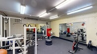 This single-room fitness facility offers a well-organized and fully equipped workout space designed to accommodate a wide range of exercise disciplines. The room is constructed with concrete block walls finished in a neutral tone, lending it a durable, utilitarian character suited for an active environment. Overhead fluorescent lighting provides consistent, bright illumination throughout the space, ensuring excellent visibility during workouts.

The flooring is covered in dark rubber matting, a practical and resilient surface ideal for heavy equipment and high-impact activities. Large wall-mounted mirrors span multiple walls, visually expanding the room and allowing users to monitor their form during exercises.

The space is thoughtfully outfitted with a diverse selection of equipment. Strength training is well-supported with a multi-station weight machine, a squat and pull-up rack, an adjustable bench, and a full rack of dumbbells and kettlebells with accompanying weight plates. A freestanding Wave Master punching bag serves as a dedicated boxing and martial arts training station.

Cardio options include a treadmill, a stationary spin bike, and a ski-stride elliptical-style trainer. A stability ball and additional accessories round out the functional training offerings. Exposed piping and ductwork run along the ceiling, reinforcing the facility's practical, no-frills aesthetic. The space is accessed through a single solid door and provides a focused, efficient environment for comprehensive fitness training.