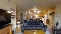 This single-floor space showcases an open-concept design that seamlessly integrates the living and kitchen areas into one cohesive environment. The living room is anchored by a large, dark leather sofa adorned with Western-themed star-embroidered throw pillows, complemented by warm wood-toned side tables and a matching coffee table. A ceiling fan with integrated lighting is centrally positioned overhead, providing both comfort and illumination to the space. The neutral-toned walls and light carpeting create a understated backdrop that highlights the Western-inspired décor, including cross wall accents and a framed mirror. The kitchen, visible from the living area, features light wood cabinetry extending to the upper walls, providing generous storage. Full-sized appliances including a white refrigerator, range, and over-the-range microwave are integrated into the layout. A dining area with a dark wood table and chairs bridges the two spaces, reinforcing the open and functional flow. The overall ambiance is casual and warm, with a cohesive rustic Western theme carried throughout the furnishings and decorative elements.