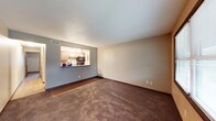 This single-floor residential space is designed with an open and functional layout that encourages seamless flow between the living and kitchen areas. The living room features wall-to-wall carpeting in a warm, earthy brown tone, complemented by dark wood baseboards that add a classic finishing touch. Neutral-toned walls in soft beige and gray create a calm, versatile backdrop throughout the space.

A defining architectural feature is the pass-through kitchen window, which opens the kitchen to the living area, enhancing the sense of openness and making the space feel larger and more connected. The kitchen is equipped with wooden cabinetry in a light honey finish, a tile mosaic backsplash, and black appliances including a built-in microwave. Overhead pendant lighting in the kitchen provides focused illumination, adding both functionality and a decorative element.

The entryway transitions from hardwood-style flooring into the carpeted living area, providing a clear and practical delineation between zones. A ceiling vent ensures climate control throughout the space. Overall, this unit presents a straightforward, well-organized layout suited for comfortable everyday living.