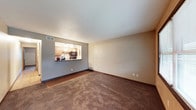 This single-floor residential space is designed with an open and functional layout that encourages seamless flow between the living and kitchen areas. The living room features wall-to-wall carpeting in a warm, earthy brown tone, complemented by dark wood baseboards that add a classic finishing touch. Neutral-toned walls in soft beige and gray create a calm, versatile backdrop throughout the space.

A defining architectural feature is the pass-through kitchen window, which opens the kitchen to the living area, enhancing the sense of openness and making the space feel larger and more connected. The kitchen is equipped with wooden cabinetry in a light honey finish, a tile mosaic backsplash, and black appliances including a built-in microwave. Overhead pendant lighting in the kitchen provides focused illumination, adding both functionality and a decorative element.

The entryway transitions from hardwood-style flooring into the carpeted living area, providing a clear and practical delineation between zones. A ceiling vent ensures climate control throughout the space. Overall, this unit presents a straightforward, well-organized layout suited for comfortable everyday living.