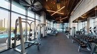 Located at 2109 S Wabash Ave in Chicago's South Loop, this contemporary fitness center is designed to deliver a high-performance workout environment with a polished, industrial-modern aesthetic. The space features soaring ceilings with exposed structural elements, dark painted ductwork, and dramatic cross-pattern linear LED lighting that lends the facility a sleek, energetic atmosphere.

Floor-to-ceiling glass walls line the perimeter, flooding the interior with abundant natural light and offering open views of the surrounding cityscape and an adjacent outdoor pool area. Large ceiling fans provide air circulation throughout the expansive floor plan, which is covered in durable rubber tile flooring suited for heavy use.

The facility is comprehensively equipped with a wide array of cardio machines, including treadmills, ellipticals, stair climbers, stationary bikes, and rowing machines positioned to take advantage of the exterior views. The strength training section features cable machines, free weights, weight benches, and plate-loaded equipment from brands including Precor and TAG. Exposed concrete columns punctuate the space, reinforcing its industrial character while defining functional zones.

A dedicated TRX Training Zone offers a wall-mounted suspension training rig alongside full-length mirrors, providing a separate area for functional and bodyweight training. The overall design balances form and function, creating a motivating and well-appointed fitness environment.