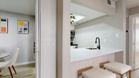 This well-designed single-floor residence showcases a clean, contemporary aesthetic throughout, with light wood-style flooring and neutral gray tones creating a cohesive and airy feel across all rooms. The open-concept kitchen is a standout feature, outfitted with crisp white cabinetry, striking marble-look quartz countertops, a matte black pull-down faucet, and a full suite of stainless steel appliances including a refrigerator, range, microwave, and dishwasher. A pass-through breakfast bar with gold-accented stools connects the kitchen to the adjacent living and hallway areas, enhancing the sense of openness. The living room benefits from a ceiling fan, vertical blinds, and a sliding door that allows natural light to fill the space. The bedroom is generously proportioned, featuring a ceiling fan, large windows with natural light exposure, and ample room for full furniture arrangements. The bathroom is finished with a white shaker-style vanity, marble-look quartz countertop, matte black fixtures, and a tub and shower surround clad in elegant marble-look tile. Throughout the residence, consistent design choices — including black hardware accents, light flooring, and textured walls — tie the space together into a polished, move-in-ready home.
