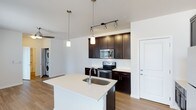 This single-floor residence offers a thoughtfully designed open-concept layout that seamlessly connects the living and kitchen areas, creating a bright and functional living environment. The kitchen serves as the centerpiece of the space, anchored by a generous island with a white quartz countertop and an undermount stainless steel sink. Dark espresso-toned cabinetry provides a bold contrast against the light countertops and neutral walls, while stainless steel appliances — including an electric range, built-in microwave, and refrigerator — add a sleek, modern finish. Overhead, a combination of track lighting and elegant pendant lights illuminates the kitchen with both practicality and style. The open layout extends naturally into the adjoining living area, enhanced by warm hardwood-style flooring throughout that ties the spaces together cohesively. A notable convenience of this residence is the in-unit laundry, with a stacked washer and dryer tucked neatly into a dedicated alcove. Clean lines, a neutral color palette, and contemporary finishes give the overall space a polished, move-in-ready ambiance suited for modern living.