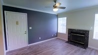 This single-story space presents a thoughtfully designed living area that combines functional layout with tasteful finishes. The room is anchored by a striking deep blue-gray accent wall that contrasts with the neutral, light-toned walls on the adjacent sides, creating a sense of depth and dimension. Crown molding runs along the ceiling perimeter, adding a classic architectural detail that elevates the overall aesthetic.

The flooring throughout is light hardwood-style planking, lending warmth and continuity to the space while remaining easy to maintain. A ceiling fan with an integrated light fixture is centrally positioned, providing both air circulation and ambient lighting. The room benefits from natural light through a window fitted with horizontal blinds, allowing for adjustable brightness.

A fireplace with a dark, granite-style surround and glass-front enclosure serves as a notable focal point along one wall, adding both visual interest and functional warmth. White baseboards frame the floors cleanly, complementing the white entry door visible on one side of the room. The open, uncluttered layout allows for versatile furniture arrangements, making the space adaptable to a variety of living needs.