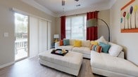 Located at 3816 Banyan Grove Lane in Virginia Beach, Virginia, this single-floor residence offers a thoughtfully designed layout with a welcoming ambiance throughout. The living room is bright and open, featuring crown molding, a ceiling fan, large windows with bold red accent curtains, and sliding glass door access, allowing generous natural light to fill the space. Light wood-style flooring flows through the main living areas, complementing the neutral wall tones.

The kitchen is well-equipped with white cabinetry, white appliances including a range and refrigerator, ample counter space, a double sink, and a peninsula with bar seating — ideal for casual dining. A built-in desk nook adds functional workspace just off the kitchen. The dining area features crown molding and chair-rail detailing, creating a refined setting for meals.

The bedrooms offer plush carpeting and distinctive accent walls — one in a vibrant teal and another in bold yellow — providing character and warmth. Each room is served by natural light from well-placed windows. The two bathrooms each include a soaking tub and shower combination, with light wood-style flooring, white vanity cabinetry, and clean, neutral finishes throughout. The overall design balances comfort and practicality in every room.