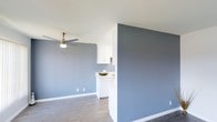 This single-floor space showcases a clean, modern aesthetic defined by cool blue-grey walls and crisp white trim that together create a calm, contemporary atmosphere. The open layout allows the living area to flow naturally into the kitchen zone, which is partially separated by a half-wall partition — a functional architectural detail that maintains visual connectivity while defining each area. The kitchen features white cabinetry and a white countertop, contributing to a bright, clean look. Wood-look flooring runs throughout, adding warmth and continuity to the space. A ceiling fan with an integrated light fixture is centrally positioned in the living area, providing both air circulation and overhead lighting. White baseboards run along the perimeter, offering a polished finish that contrasts neatly with the blue-grey walls. The overall design is minimal and well-coordinated, with a neutral palette that offers flexibility for various furnishing styles. The layout is efficient and practical, making effective use of the available footprint on a single level.