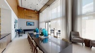 This thoughtfully designed single-floor amenity space showcases an impressive array of social and recreational areas unified by a cohesive contemporary aesthetic. The architecture is defined by dramatic double-height ceilings that amplify the sense of openness throughout, while floor-to-ceiling glass walls and large double-door entries flood the interiors with natural light and provide views of surrounding tropical landscaping.

The main gathering area features a generously sized conference-style dining table accommodating numerous guests, adorned with sculptural blue glass décor and suspended wire-frame pendant lights. Adjacent to this is a lounge area anchored by warm wood-paneled walls inset with lush living moss panels, complemented by a curated mix of seating including a light blue sofa, gray armchairs, and a bold yellow bench.

A standout feature is the three-dimensional geometric accent wall composed of gold, silver, and navy faceted tiles, serving as a dramatic focal point near a built-in service counter. A dedicated game area includes a full shuffleboard table beneath a wall-mounted television. A casual café-style seating nook with blue booth seating, warm wood-plank walls, and teal pendant lights rounds out the space.