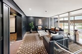 Located at 265 Reynolds Street in Oakville, Ontario, this building lobby presents a refined and sophisticated entrance experience. The space is defined by dramatic dark-stained wood panel wainscoting that lines the walls throughout, creating a cohesive and upscale aesthetic. Recessed lighting illuminates the area with a warm, inviting glow, complementing the clean white ceilings above.

The central lounge area is thoughtfully furnished with sleek black leather seating — including sofas and armchairs — arranged around a contemporary glass-topped coffee table, all set atop a richly patterned geometric area rug. A large-scale equestrian portrait serves as a striking focal point on the feature wall, adding an artistic touch. Lush indoor plants provide natural accents throughout the space.

Floor-to-ceiling windows and glazed walls on one side flood the lobby with abundant natural light and offer open views of the surrounding greenery and streetscape, creating a bright contrast to the darker interior finishes. The elevator corridor continues the dark panelled design language, with patterned carpet runner flooring and dark-stained doors that maintain a consistent, polished character. Together, these elements create a lobby that is both functional and impressively designed.