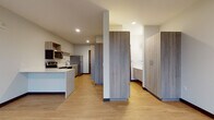 Located at 9150 W Watertown Plank Rd in Wauwatosa, Wisconsin, this modern single-floor unit showcases a clean, contemporary design with consistent light wood-tone flooring and neutral walls throughout. The open-concept kitchen and living area serves as the heart of the space, featuring warm wood-grain cabinetry, crisp white countertops, and a full suite of stainless steel appliances including an electric range, built-in microwave, dishwasher, and refrigerator. A peninsula counter provides additional workspace and a natural division between the kitchen and the open living area. Natural light flows in through a set of large windows, offering views of the surrounding outdoor landscape and lending the living space an airy, open feel. A wall-mounted split system unit provides climate control. The bedroom is generously sized, with the same light wood flooring and large windows that frame outdoor views. Tall wood-grain storage cabinets provide ample built-in storage. The bathroom features a clean, minimalist design with a wood-grain vanity, white countertop, and a dedicated walk-in shower with brushed nickel fixtures. Recessed lighting throughout the unit adds a polished, modern finish to the overall aesthetic.
