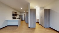 Located at 9150 W Watertown Plank Rd in Wauwatosa, Wisconsin, this modern single-floor unit showcases a clean, contemporary design with consistent light wood-tone flooring and neutral walls throughout. The open-concept kitchen and living area serves as the heart of the space, featuring warm wood-grain cabinetry, crisp white countertops, and a full suite of stainless steel appliances including an electric range, built-in microwave, dishwasher, and refrigerator. A peninsula counter provides additional workspace and a natural division between the kitchen and the open living area. Natural light flows in through a set of large windows, offering views of the surrounding outdoor landscape and lending the living space an airy, open feel. A wall-mounted split system unit provides climate control. The bedroom is generously sized, with the same light wood flooring and large windows that frame outdoor views. Tall wood-grain storage cabinets provide ample built-in storage. The bathroom features a clean, minimalist design with a wood-grain vanity, white countertop, and a dedicated walk-in shower with brushed nickel fixtures. Recessed lighting throughout the unit adds a polished, modern finish to the overall aesthetic.