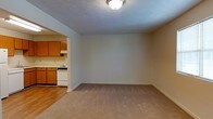 This single-floor residential unit offers a straightforward and functional layout designed for comfortable everyday living. The main living area is finished with wall-to-wall carpeting in a neutral beige tone, creating a warm and understated atmosphere. The walls are painted in a soft, light color that keeps the space feeling bright and clean, complemented by a flush-mount ceiling light fixture overhead. A textured ceiling adds a subtle architectural detail throughout the space.

An open passageway connects the living area directly to the kitchen, which features warm-toned wooden cabinetry providing ample storage. The kitchen is equipped with a white range and oven, as well as a dishwasher, offering practical functionality for daily meal preparation. The kitchen floor transitions to hardwood-style flooring, creating a clear visual distinction between the two spaces.

The open connection between the living area and kitchen promotes an easy flow and a sense of cohesion throughout the unit. The layout is clean and uncluttered, offering a blank canvas that can be easily personalized. Overall, the space presents a modest yet comfortable environment well-suited for practical residential living.