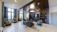 This single-level lobby presents a refined, contemporary aesthetic that immediately establishes an atmosphere of sophistication. The space is anchored by a striking dark wood feature wall, which houses a large flat-screen television and integrated shelving with decorative accents and ambient uplighting. A suspended square pendant light draws the eye upward, complementing the recessed ceiling lighting and the contrasting dark ceiling panel that defines the central seating zone.

The seating area is thoughtfully arranged with a substantial gray sectional sofa, chrome-framed glass coffee tables, and a sleek round pedestal side table. A teal accent chair adds a pop of color, echoing the floor-length teal drapery panels that frame the large windows along the perimeter walls. These windows allow generous natural light to filter into the space, brightening the light-toned tile flooring.

Additional seating, including a classic wingback chair with a vibrant accent pillow, is positioned near the windowed wall, offering a quieter retreat. Potted greenery and carefully placed decorative objects contribute to the polished yet inviting ambiance of this well-appointed lobby.