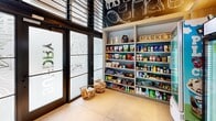 This self-service market is a compact yet well-appointed retail space designed to provide convenient access to everyday essentials. Entry is made through striking floor-to-ceiling glass doors adorned with frosted tropical leaf motifs, framed in sleek black metal — immediately establishing a modern aesthetic. The interior is defined by a bold, large-scale chalkboard-style wall mural depicting illustrated grocery items such as chips, moisturizer, detergent, and more, lending the space a playful yet informative character.

The retail area is efficiently organized, featuring multiple glass-door refrigeration units stocked with beverages, dairy, and chilled snacks, alongside a dedicated freezer unit. Built-in open shelving units line the walls, offering a wide variety of packaged goods including pantry staples, snacks, condiments, and household products. A self-checkout kiosk provides a seamless, cashless purchasing experience. Woven basket displays near the entrance add a warm, approachable touch to the otherwise modern interior.

Large windows allow natural light to filter in, complementing the light-toned tile flooring that runs throughout. The overall design balances functionality with visual interest, making this market a convenient and inviting stop for daily needs.